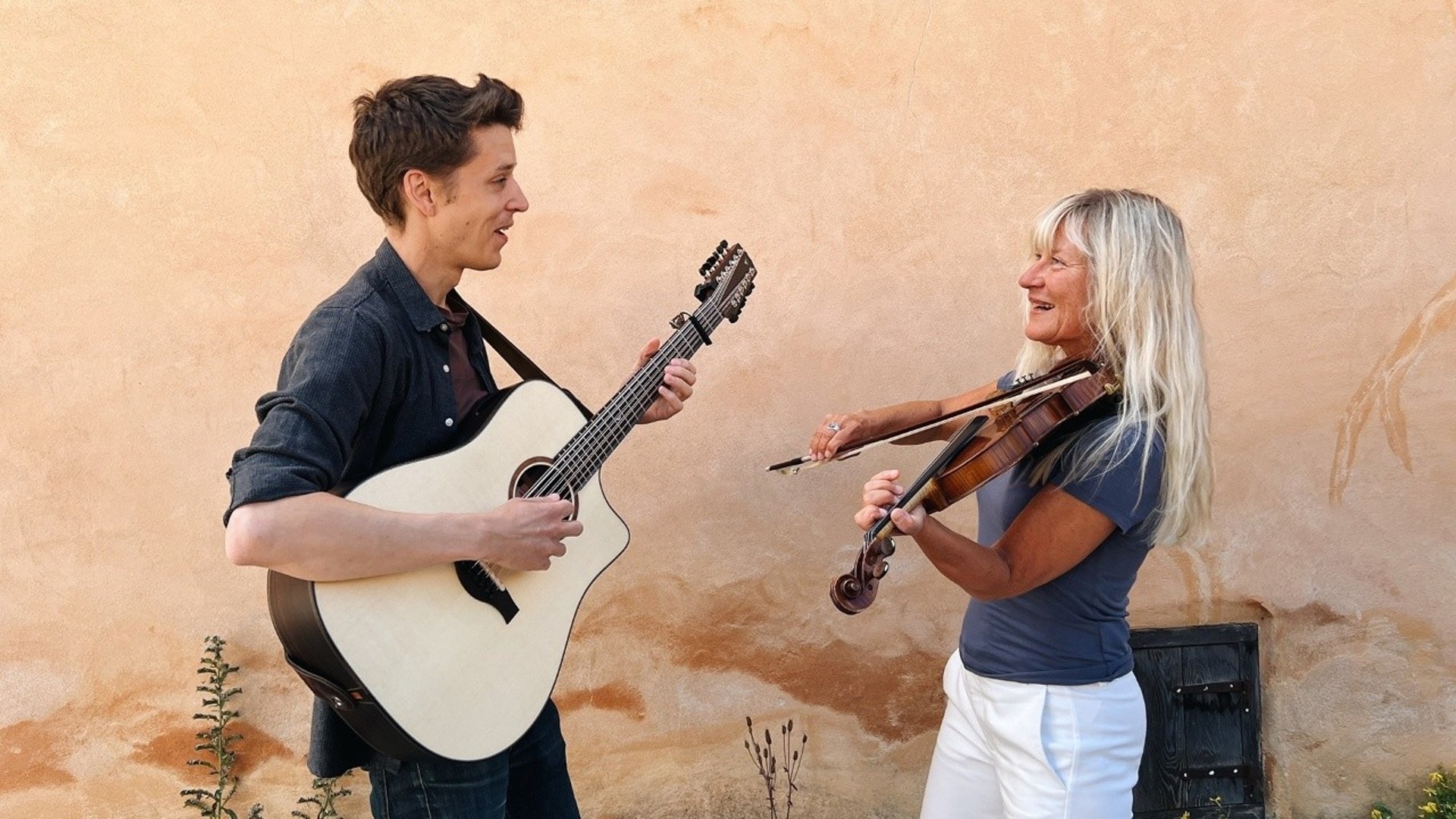 Bild på Simon med gitarr och Lena med fiol som står vända mot varandra framför en ljusgul vägg .