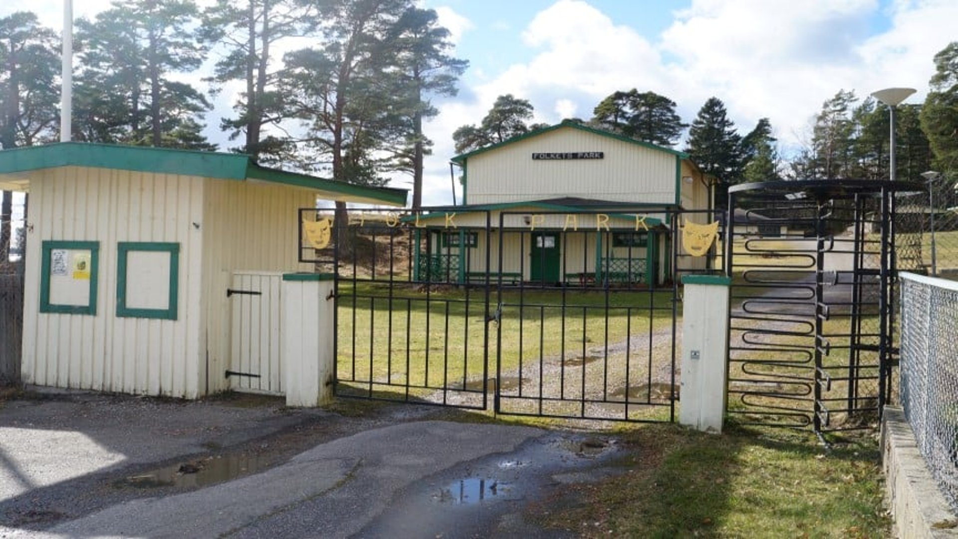 Entrén till Nävekvarns Folkets park, med en metallgrind och en "utgångssnurra" samt parkens gula hus med gröna detaljer och grusvägen som leder vidare in i parken.
