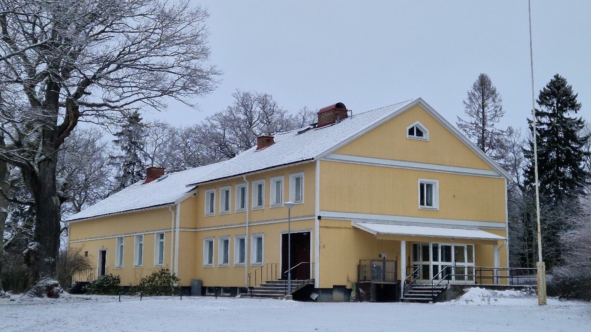 Folkets Hus i Vrena. En gul tvåvåningsbyggnad med vita knutar och snötäckt tak i vintermiljö. Framför huset ligger en snötäckt gårdsplan omgiven av träd.