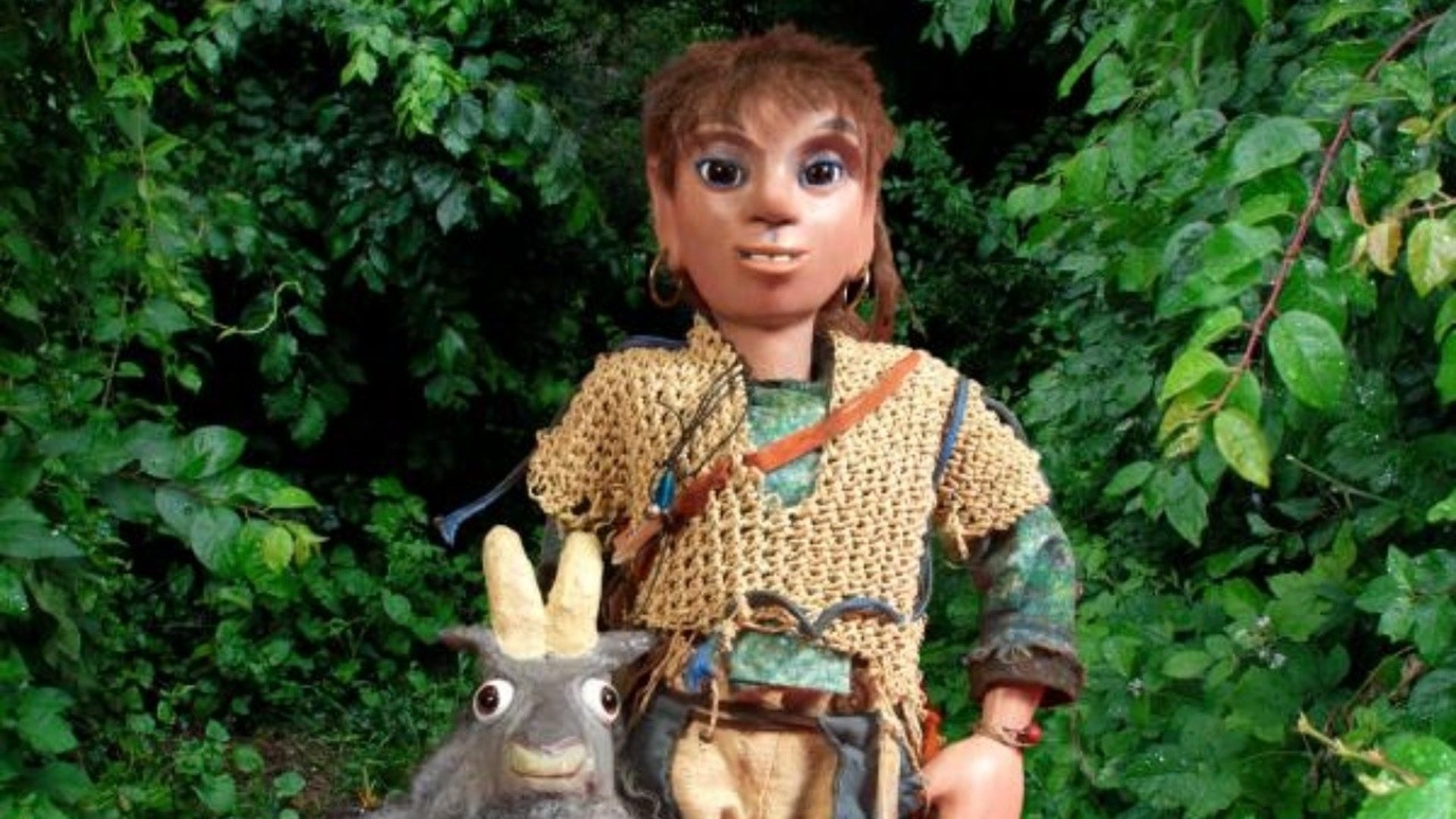 En docka av en pojke som står bredvid en docka av en get. I bakgrunden är det gröna buskar.