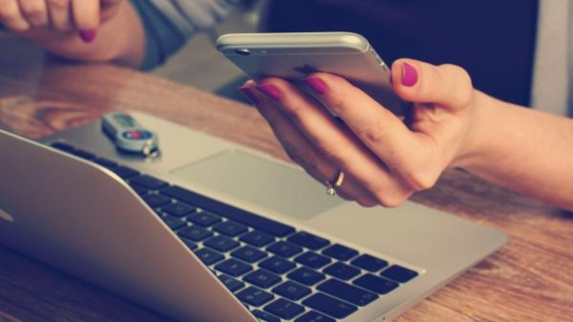 En person sitter framför en laptop och har en mobiltelefon i handen.