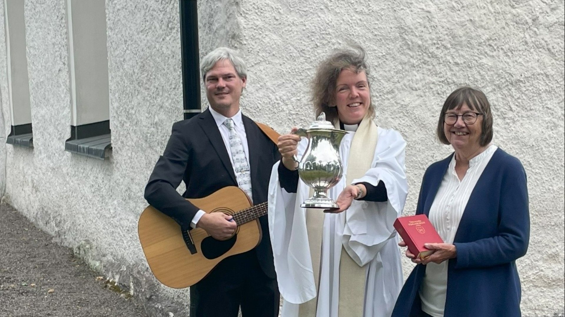 Prästen håller i dopkannan. Musikern spelar gitarr. Kyrkvärden håller i psalmboken.