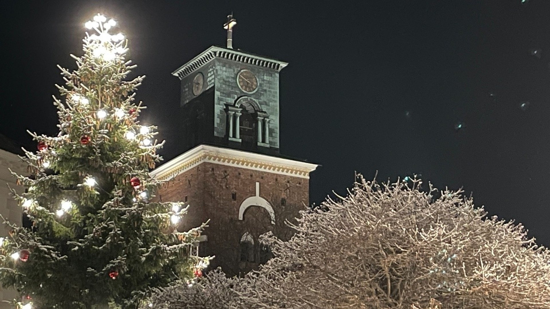 S:t Nicolai kyrktorn kvällstid med julgran och snöiga träd i förgrunden.