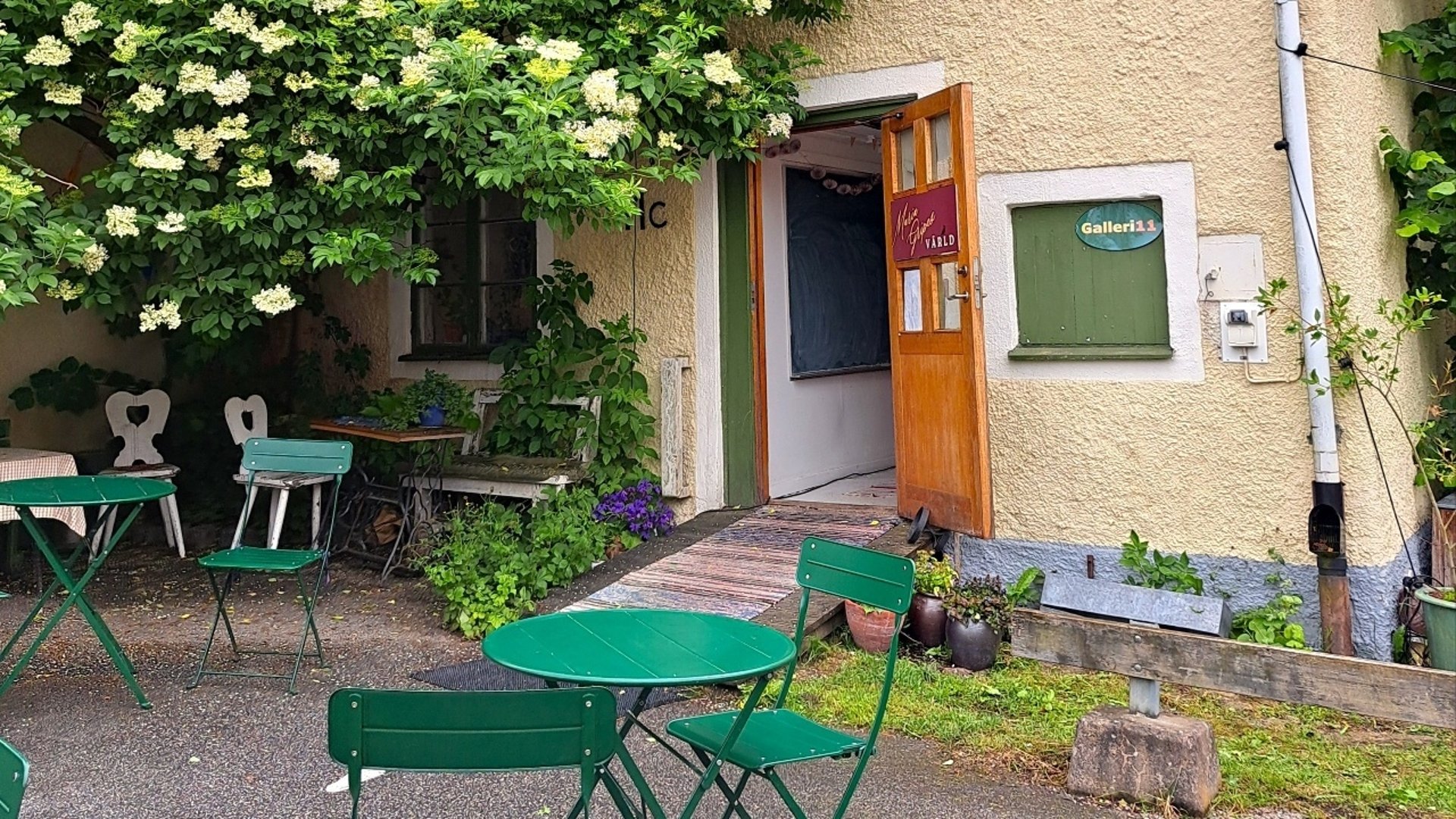 Gult stenhus med öppen trädörr. Framför dörren gröna stolar och bord. 
