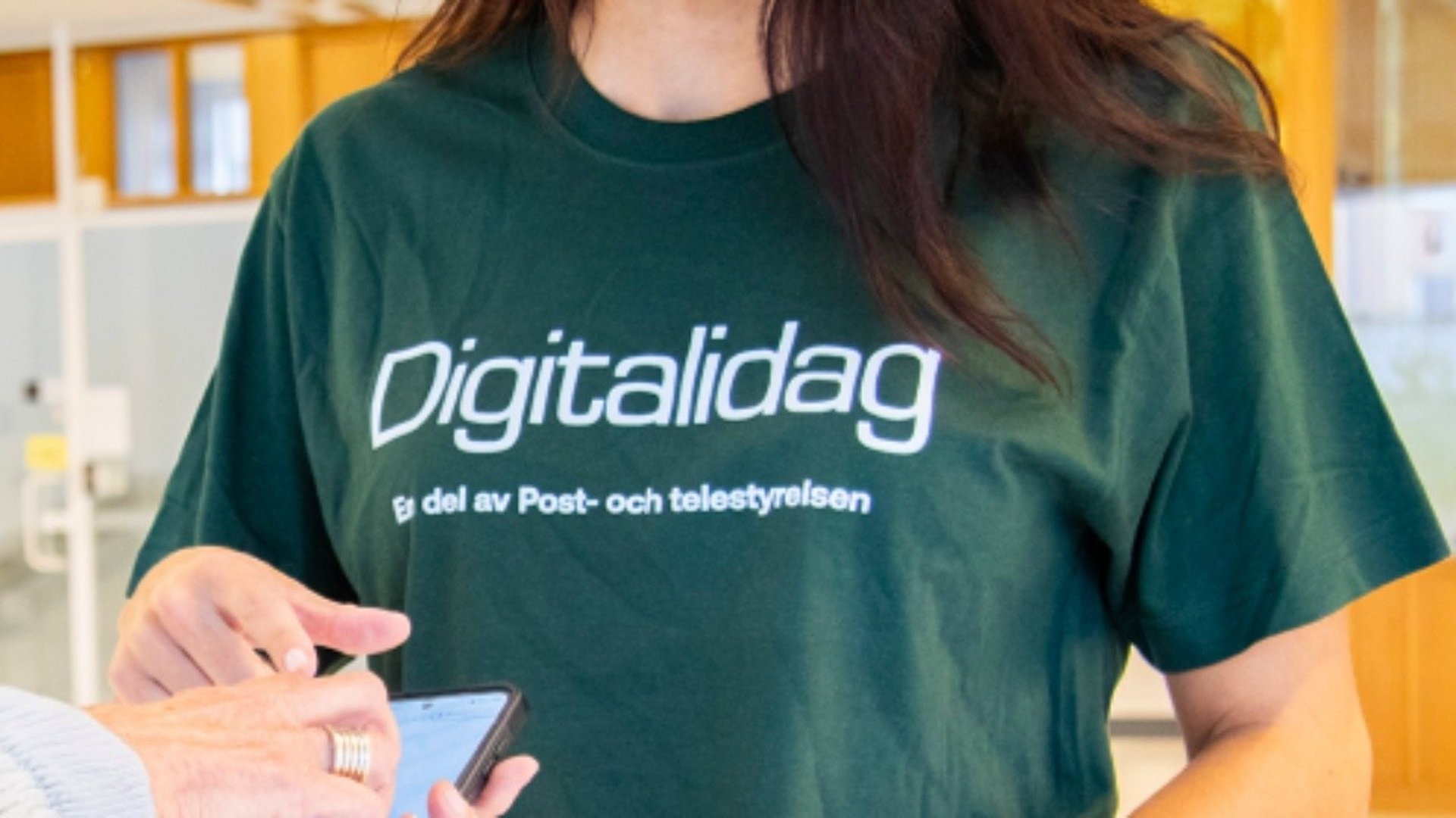 Grön t-shirt där det står Digitalidag. En person håller i en mobil och pratar med en annan person som hjälper till.