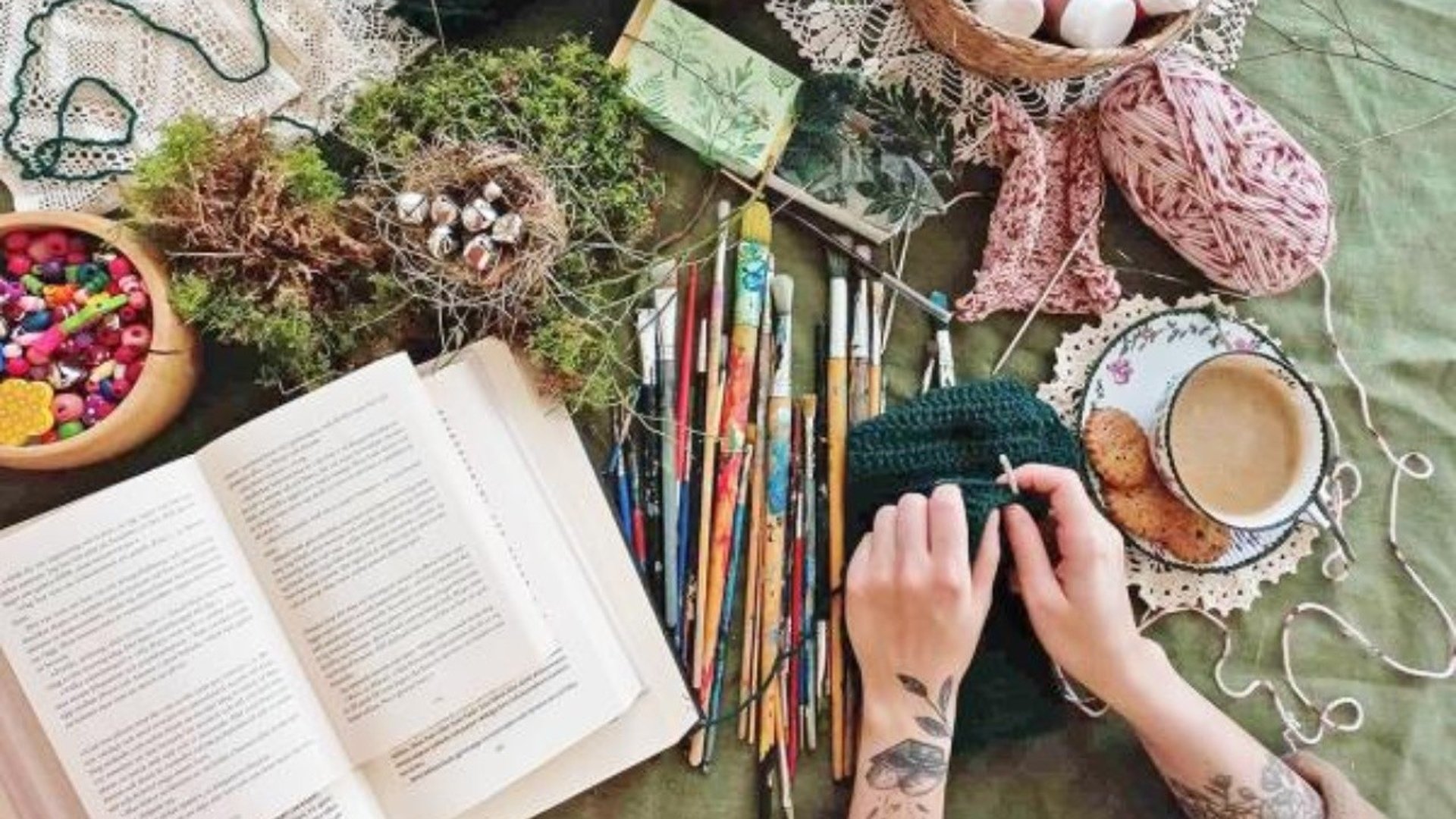 Ett bord med grön duk. Penslar, verktyg, en bok och en kaffekopp ligger på bordet.