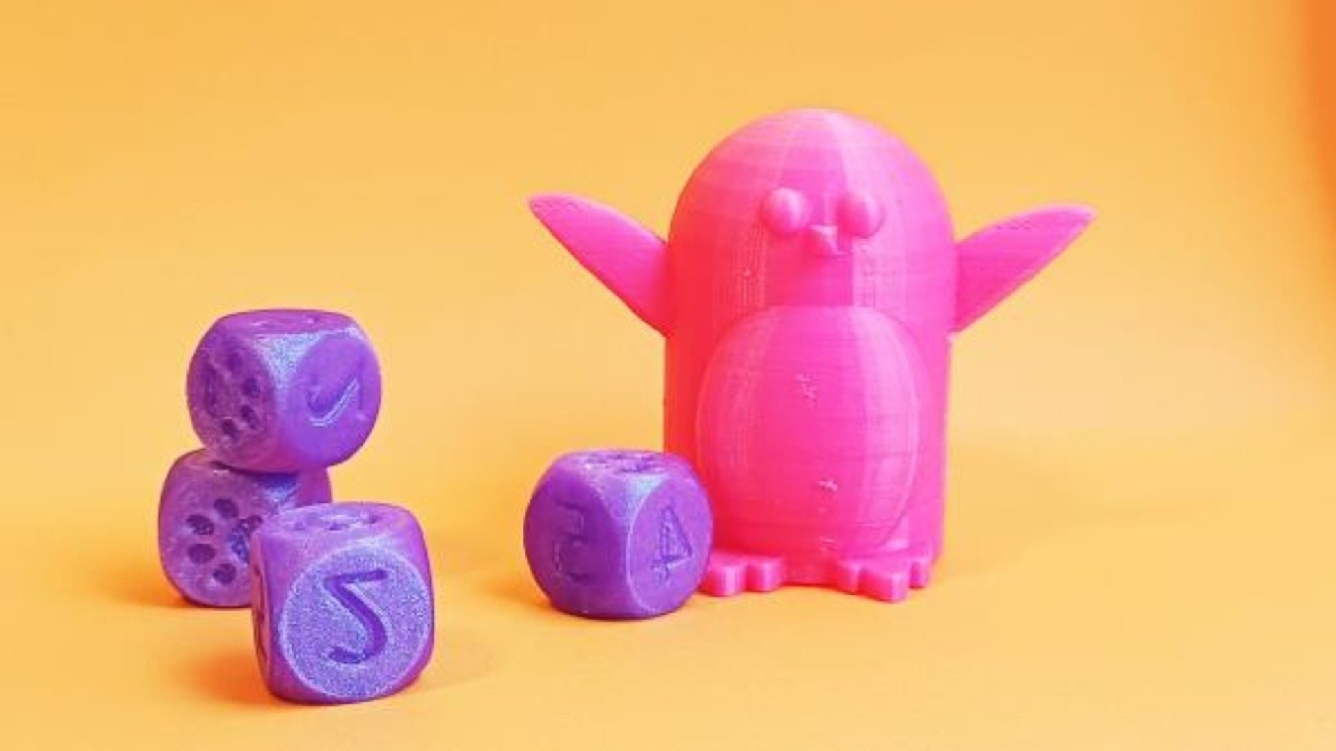 Fyra lila tärningar och en pingvin som är rosa. De är 3D-printade. Bakgrunden är orange.