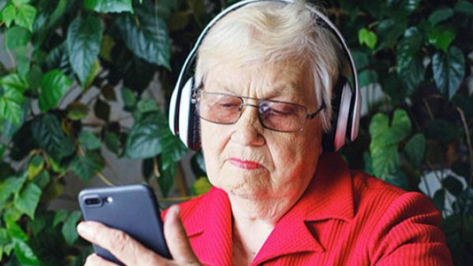 Gammal kvinna i röd skjorta sitter med hörlurar på och har sin mobil i handen.