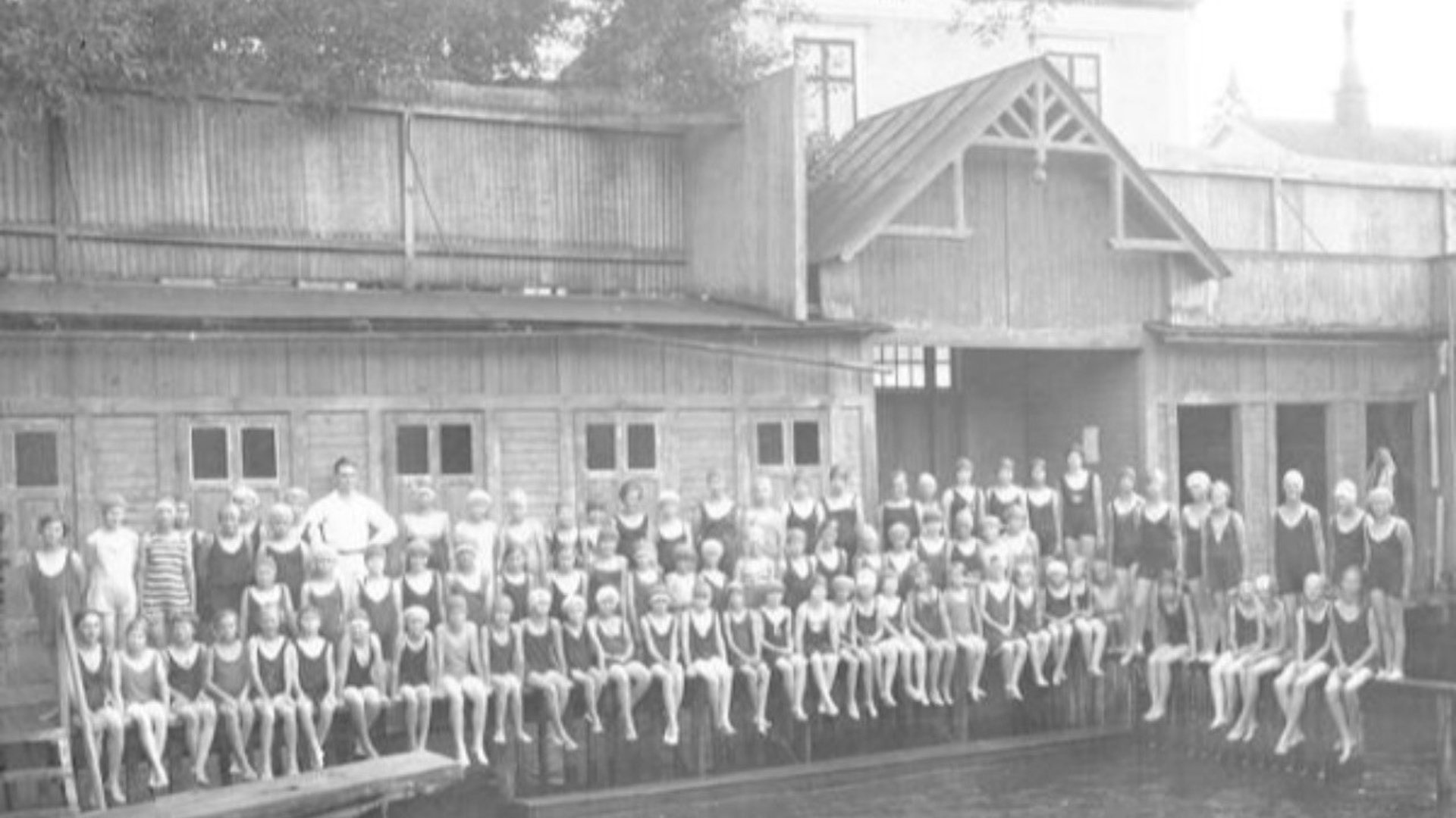 Ett gammalt svart/vitt foto på en samling barn som sitter vid ett kallbadhus.