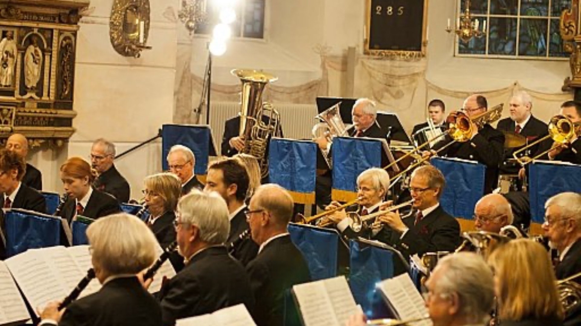 Musikkår som sitter i kyrkan och spelar på klarinetter, trumpeter och tromboner.