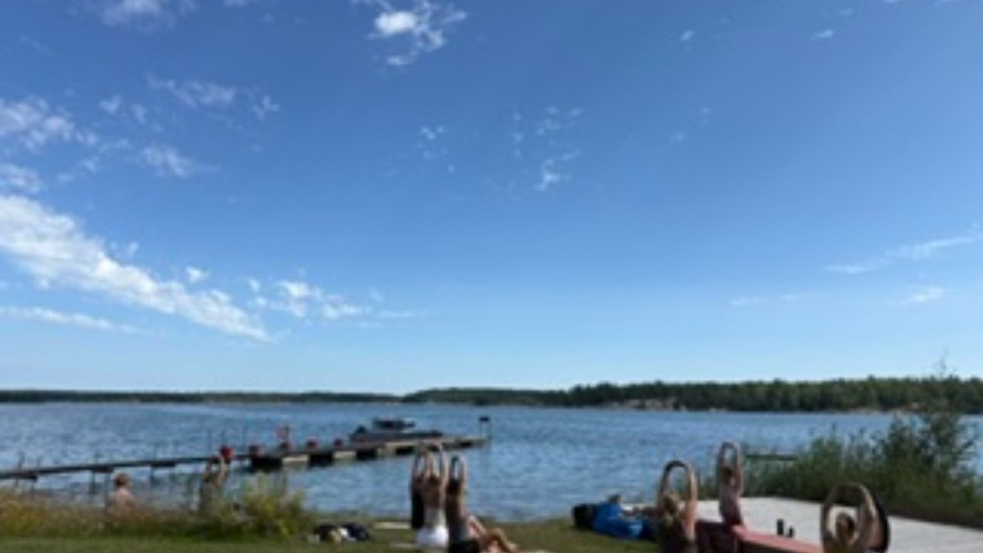 Personer som gör yoga på en strand. Hav, land och en brygga syns i mellangrunden och i bakgrunden.