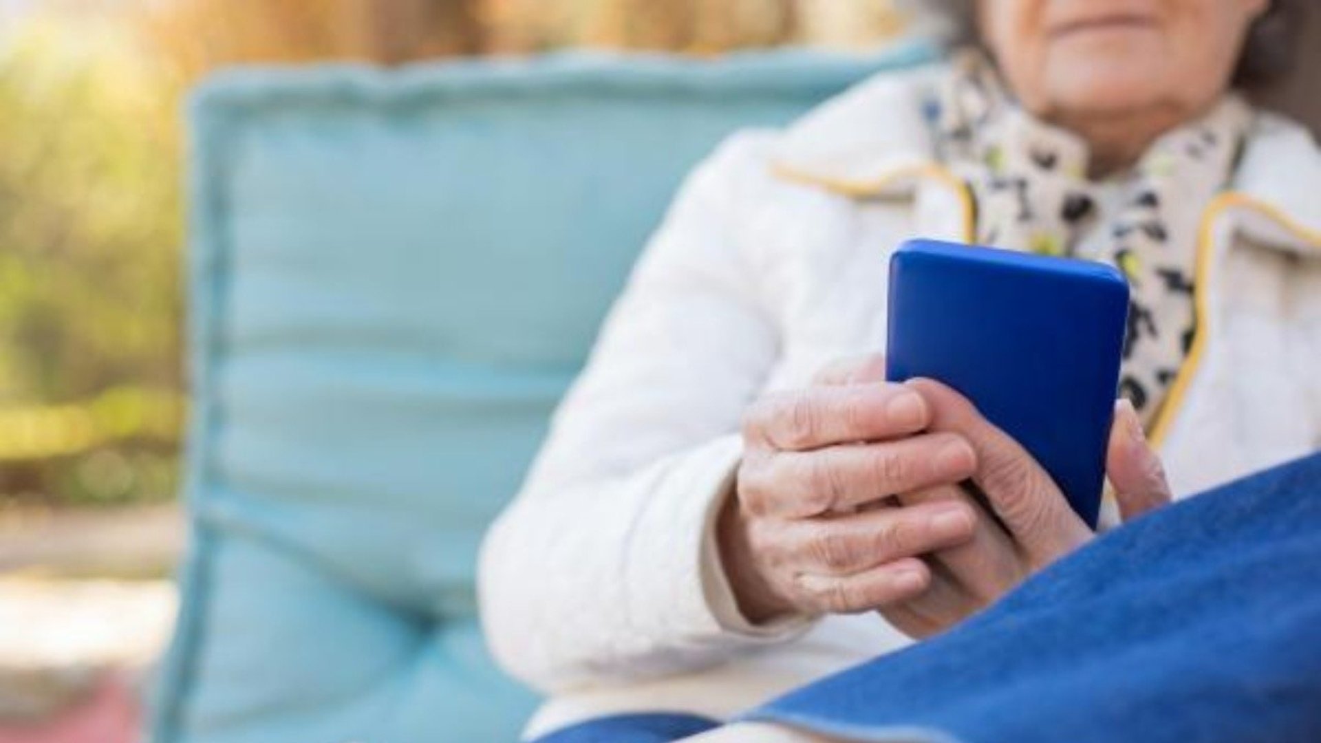 En äldre kvinna sitter i en blå fåtölj utomhus med en blå mobiltelefon i handen.