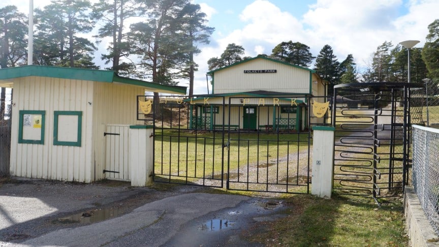 Entrén till Nävekvarns Folkets park, med en metallgrind och en "utgångssnurra" samt parkens gula hus med gröna detaljer och grusvägen som leder vidare in i parken.