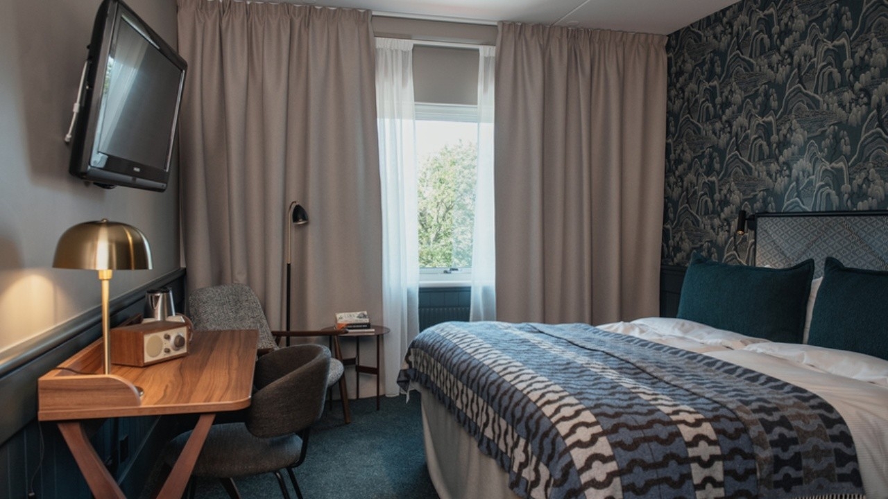 Modernt hotellrum inrett i blått med dubbelsäng, skrivbord och TV. 