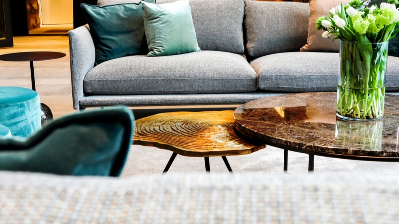 Bild på grå soffa med färgglada kuddar med soffbord som har en stor vas med blommor i vita färger på. 