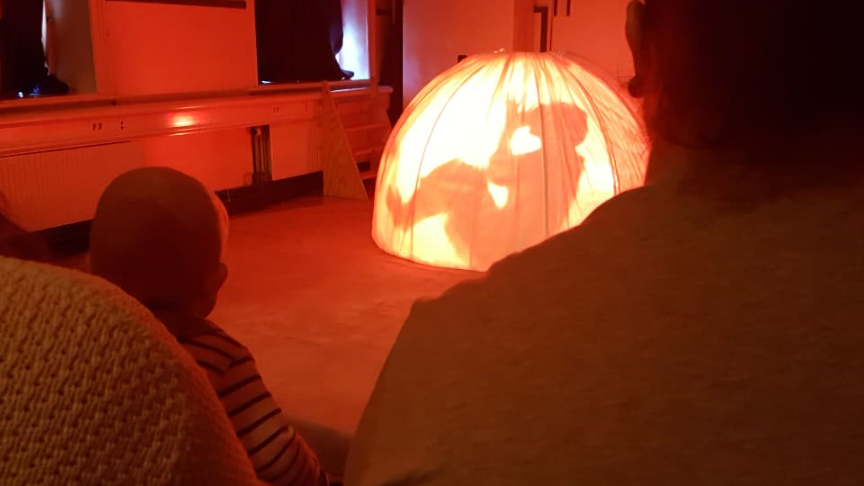 Bild ifrån Barnkulturcentrum. Man ser skuggan av en person i ett upplyst tält. Det sitter publik framför.