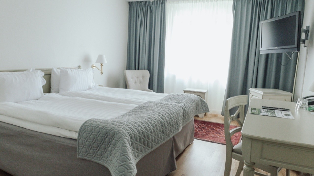 Hotellrum med dubbelsäng, vita väggar, blå detaljer, skrivbord och fönster