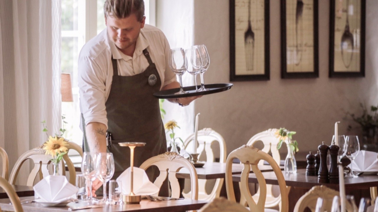 Restaurangservering där kypare ställer ned ett vinglas på bord med solrosor och naturligt ljus.